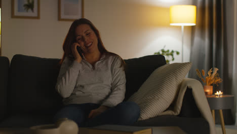 Primer-Plano-De-Una-Mujer-Sonriente-Sentada-En-Un-Sofá-En-Casa-Por-La-Noche-Hablando-Por-Teléfono-Móvil-Filmada-En-Tiempo-Real